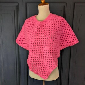 Peut inclure: Un poncho rose vif en crochet à mailles ajourées, présenté sur un mannequin. Le poncho a une encolure ronde et des manches courtes. Le motif au crochet crée un design texturé et aéré. Le vêtement est de couleur unie et le style est décontracté.