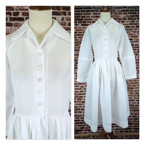 Puede incluir: Un vestido blanco con mangas largas y cuello camisero. El vestido presenta una parte delantera abotonada y una falda plisada. La tela tiene un sutil patrón texturizado. El vestido se exhibe en un maniquí contra una pared de ladrillos.