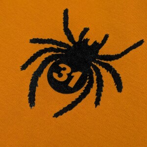May include: Close-up of orange fabric featuring a black spider design. The spider has a circular body with the number 31 inside. The spider's legs are long and textured. The design is centered.