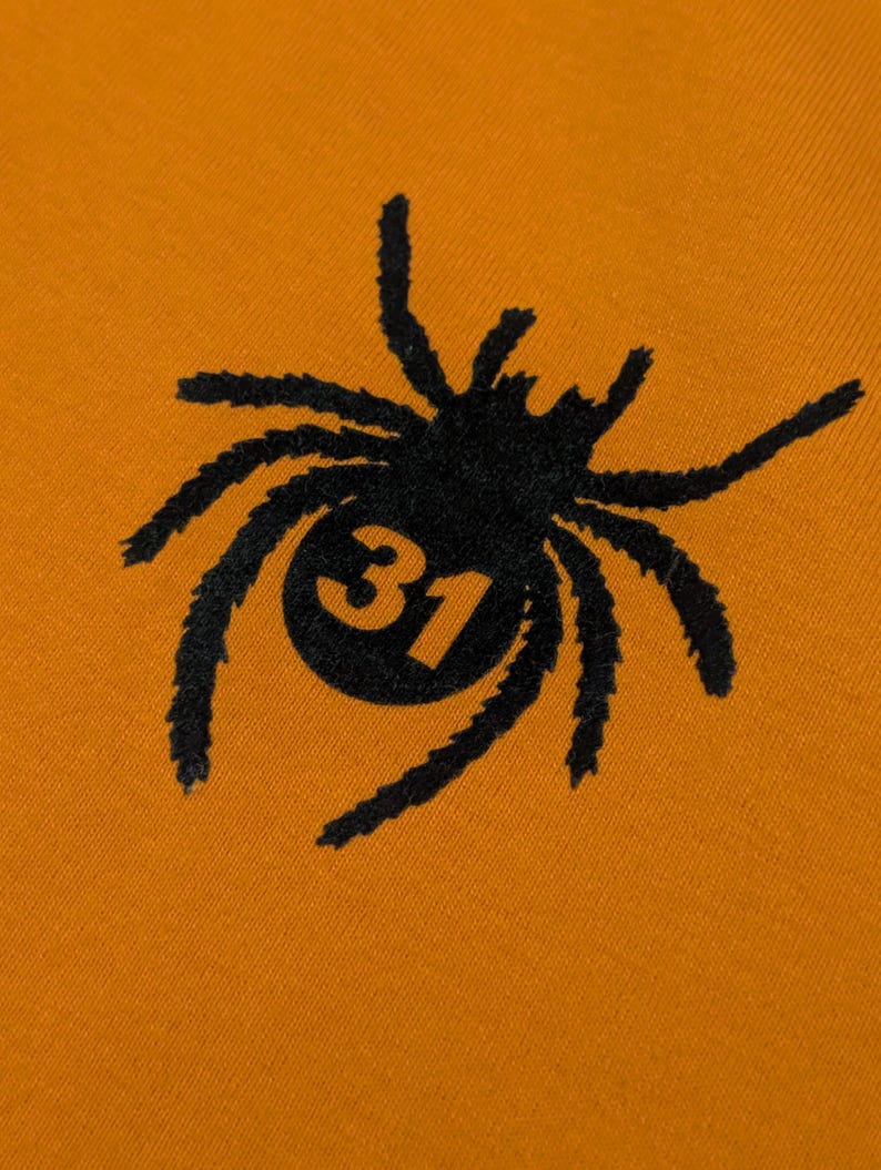 May include: Close-up of orange fabric featuring a black spider design. The spider has a circular body with the number 31 inside. The spider's legs are long and textured. The design is centered.