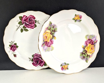 May include: Two white ceramic plates with a scalloped edge. Each plate features a floral design with purple and yellow roses, green leaves, and gold trim. The plates are set against a dark background.