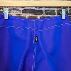 Puede incluir: Primer plano de un pantal&oacute;n corto azul vibrante colgado de una percha de madera. El pantal&oacute;n corto tiene costuras blancas y una peque&ntilde;a etiqueta negra con un logotipo blanco. La tela tiene un ligero brillo.