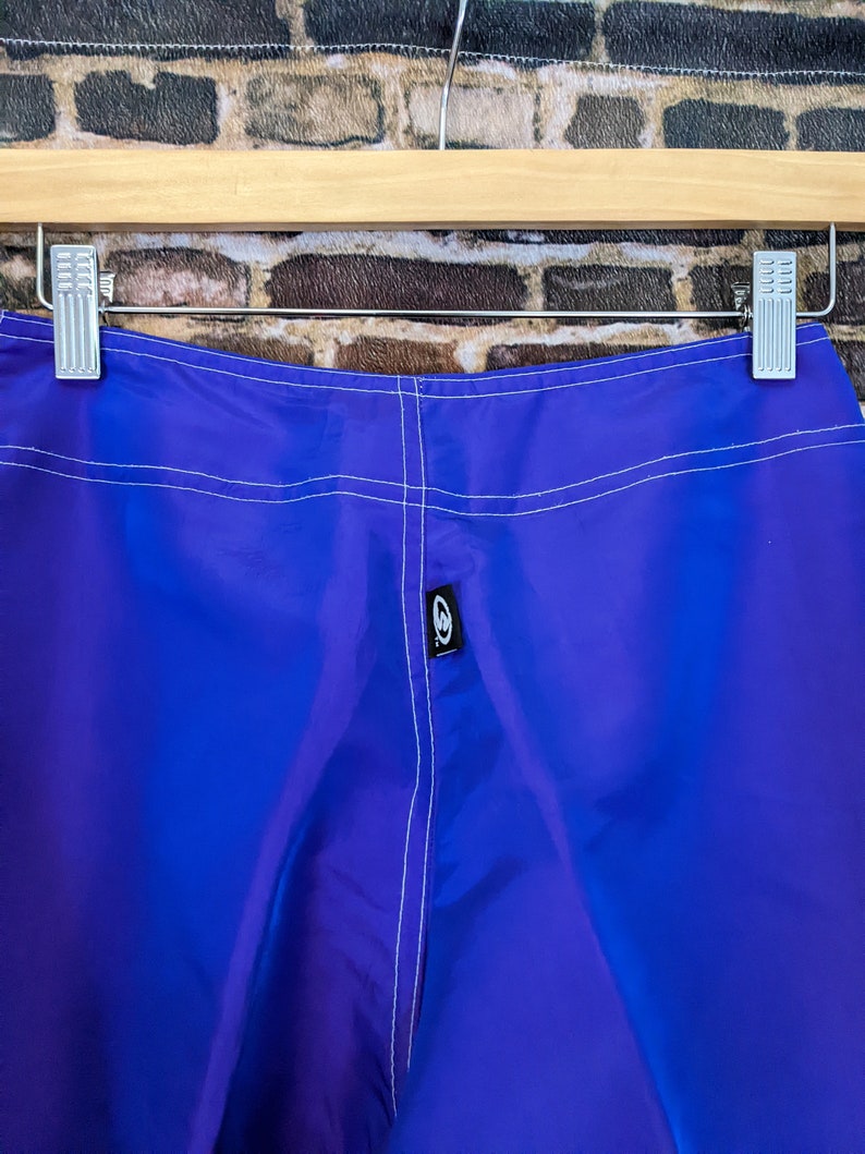 Puede incluir: Primer plano de un pantal&oacute;n corto azul vibrante colgado de una percha de madera. El pantal&oacute;n corto tiene costuras blancas y una peque&ntilde;a etiqueta negra con un logotipo blanco. La tela tiene un ligero brillo.