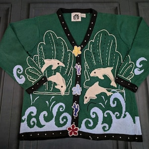 Könnte beinhalten: Grüne Strickjacke mit V-Ausschnitt, verziert mit Delfinen und Ozeanwellen. Der Pullover hat schwarze Verzierungen und dekorative Knöpfe. Die Ärmel und der Körper sind mit gestickten Delfinen, Wellen und floralen Akzenten verziert.