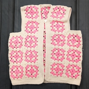 May include: A cream-colored, crocheted vest featuring a granny square design with pink floral patterns. The vest has a button closure and a sleeveless design. The garment is handmade and has a vintage aesthetic.