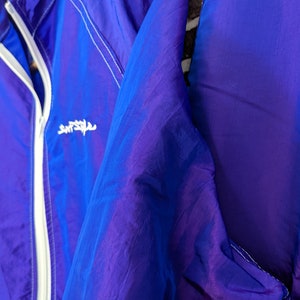Puede incluir: Chaqueta de nailon azul y morado vibrante con cremallera y ribetes blancos. La chaqueta tiene un cord&oacute;n en la parte inferior y un peque&ntilde;o logotipo blanco en el pecho. La tela tiene una calidad brillante e iridiscente.