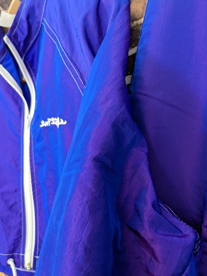 Puede incluir: Chaqueta de nailon azul y morado vibrante con cremallera y ribetes blancos. La chaqueta tiene un cord&oacute;n en la parte inferior y un peque&ntilde;o logotipo blanco en el pecho. La tela tiene una calidad brillante e iridiscente.