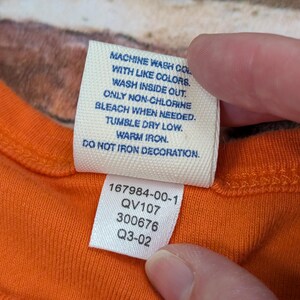 May include: Close-up of an orange garment tag with care instructions and product codes. The tag reads: Machine wash with like colours, wash inside out, only non-chlorine bleach when needed, tumble dry low, warm iron, do not iron decoration.
