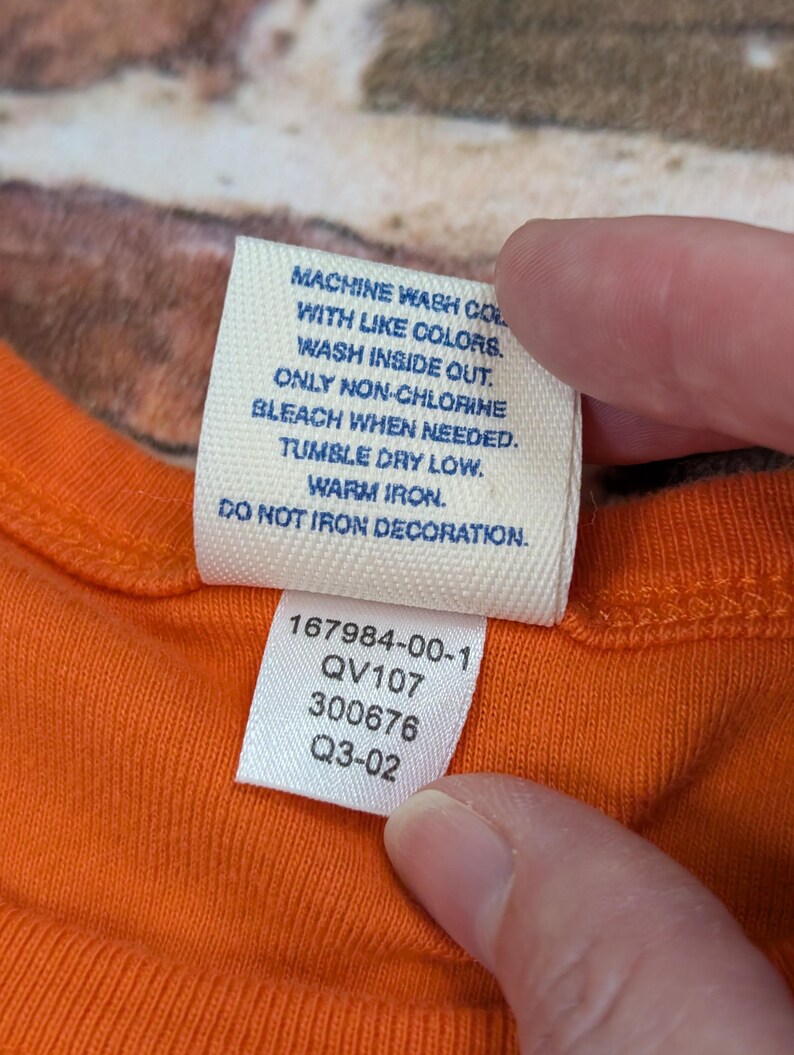 May include: Close-up of an orange garment tag with care instructions and product codes. The tag reads: Machine wash with like colours, wash inside out, only non-chlorine bleach when needed, tumble dry low, warm iron, do not iron decoration.