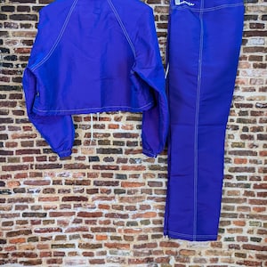 Puede incluir: Conjunto deportivo de dos piezas en morado vibrante. Incluye una chaqueta corta con cord&oacute;n y pantalones a juego. Ambas prendas presentan costuras blancas y se exhiben sobre un fondo de pared de ladrillo.