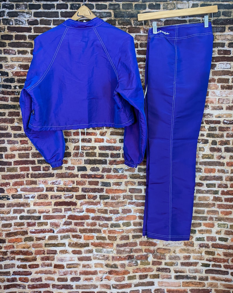 Puede incluir: Conjunto deportivo de dos piezas en morado vibrante. Incluye una chaqueta corta con cord&oacute;n y pantalones a juego. Ambas prendas presentan costuras blancas y se exhiben sobre un fondo de pared de ladrillo.