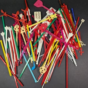 May include: A collection of vintage cocktail picks in various colors, shapes, and designs. The picks include red, yellow, green, and white, with decorative tops such as birds, musical notes, and abstract shapes. Some picks have the text "JAMES TAVERN".