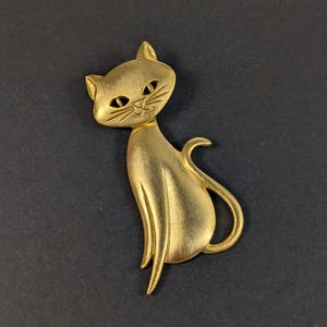 May include: A gold-colored cat brooch with a sitting pose. The brooch features a detailed design of a cat's face, body, and tail. The cat's eyes and whiskers are visible. The brooch is made of metal and has a brushed finish.