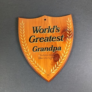 May include: Wooden shield-shaped plaque with the text "World's Greatest Grandpa" in black and gold lettering. The plaque has a gold laurel wreath design and the text "Forbidden Caverns Great Smoky Mins" in a smaller font. The plaque is made of wood.