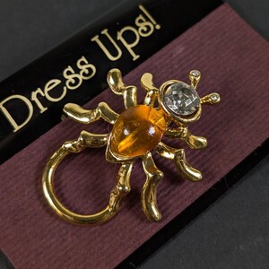 May include: A gold-toned insect brooch with an amber-colored body and a clear, faceted head. The brooch has six legs and a circular tail. The text "Dress Ups!" is visible in gold lettering on a black background.