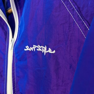 Puede incluir: Primer plano de una chaqueta cortavientos morada y azul. La chaqueta tiene una cremallera blanca y detalles de costura blancos. El texto "Surf Style" est&aacute; bordado en blanco en la parte delantera. La chaqueta es de material ligero y brillante.