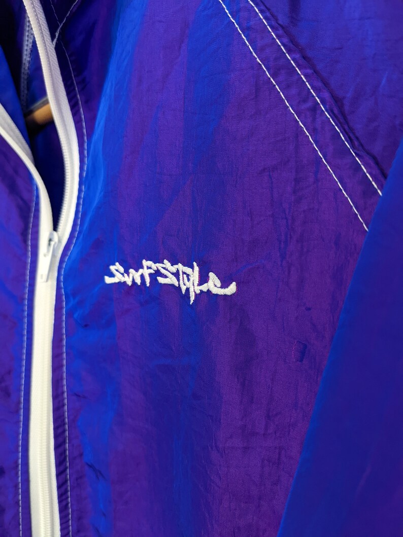Puede incluir: Primer plano de una chaqueta cortavientos morada y azul. La chaqueta tiene una cremallera blanca y detalles de costura blancos. El texto "Surf Style" est&aacute; bordado en blanco en la parte delantera. La chaqueta es de material ligero y brillante.