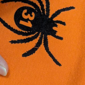 May include: Close-up of orange fabric featuring a black spider graphic. The spider has eight legs and a circular body with the number 31 inside. The fabric is a solid orange colour.