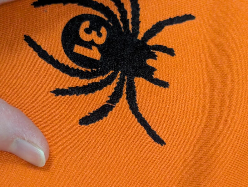 May include: Close-up of orange fabric featuring a black spider graphic. The spider has eight legs and a circular body with the number 31 inside. The fabric is a solid orange colour.