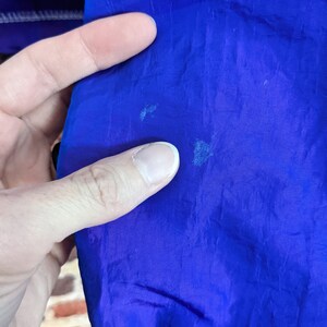 Puede incluir: Primer plano de una tela azul vibrante, posiblemente parte de una chaqueta o prenda de abrigo. El material tiene una textura suave y muestra algunas decoloraciones menores. Una mano toca la tela, resaltando el color y la textura.