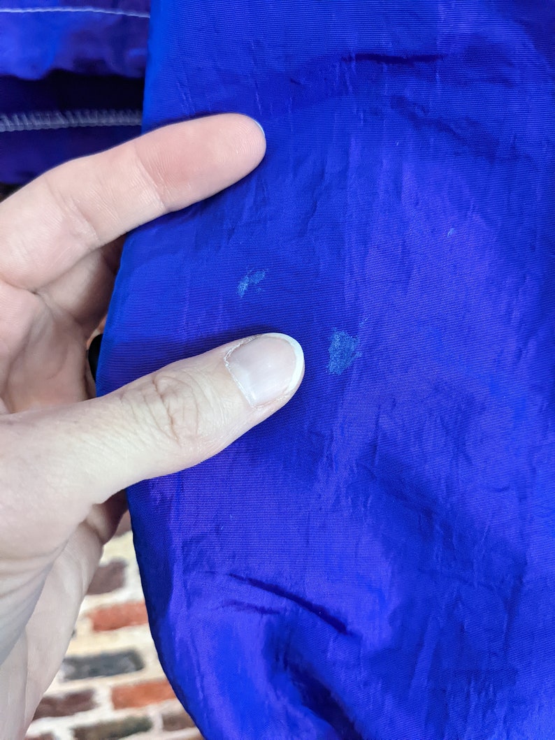 Puede incluir: Primer plano de una tela azul vibrante, posiblemente parte de una chaqueta o prenda de abrigo. El material tiene una textura suave y muestra algunas decoloraciones menores. Una mano toca la tela, resaltando el color y la textura.