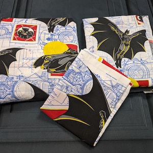 May include: A set of folded fabric featuring a Batman theme with blue technical blueprint lines on a white background. The print includes illustrations of Batman in a black cape, the Batmobile, and Catwoman, accented with yellow and red shapes.