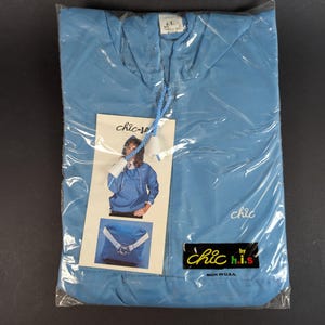 Puede incluir: Una chaqueta cortavientos azul claro nueva en un embalaje de plástico transparente. La chaqueta tiene un cuello con cordón y una pequeña etiqueta que dice "chic-j.s." y "Made in U.S.A."