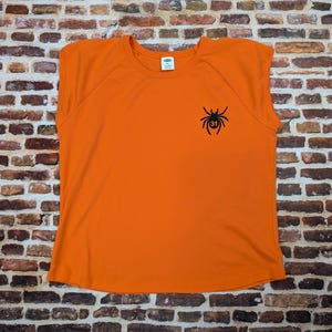 May include: A vibrant orange t-shirt featuring a black spider design with the number 31. The top has short sleeves and a crew neck. The shirt is displayed against a brick wall background.