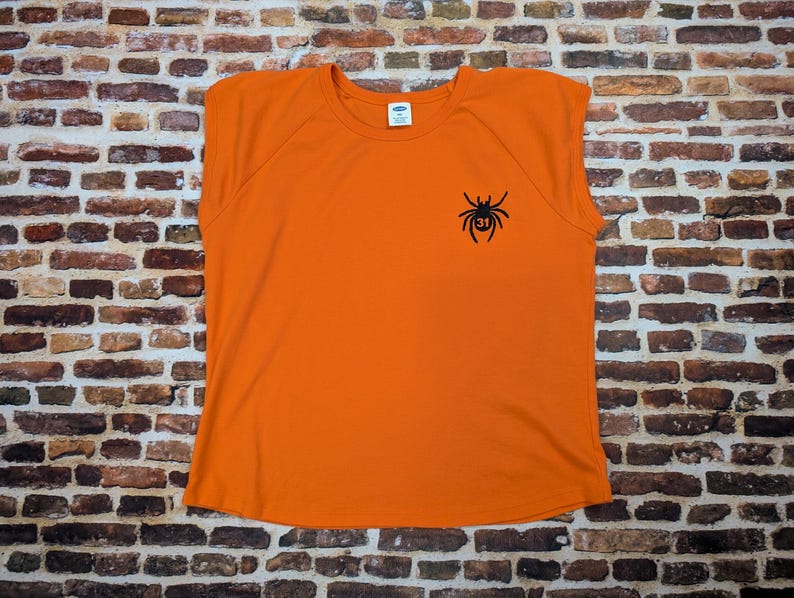 May include: A vibrant orange t-shirt featuring a black spider design with the number 31. The top has short sleeves and a crew neck. The shirt is displayed against a brick wall background.