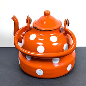 Op de afbeelding: Oranje geëmailleerde theepot met witte stippen. De theepot heeft een gebogen tuit, een deksel met een kleine knop en een grote handgreep. De handgreep en tuit zijn bevestigd met metalen scharnieren. De theepot staat op een donkere ondergrond.