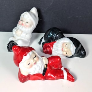 May include: Three ceramic figurines of sleeping figures with white beards and pointed hats. One figure wears a solid white outfit, one wears a black outfit, and one wears a red outfit with a black belt. Each figure has closed eyes and a serene expression.