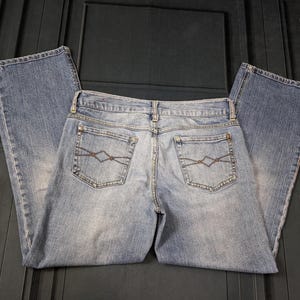 May include: Light blue denim jeans with a classic five-pocket design. The jeans feature a faded wash and decorative stitching on the back pockets. The jeans are laid flat, showcasing the back view.