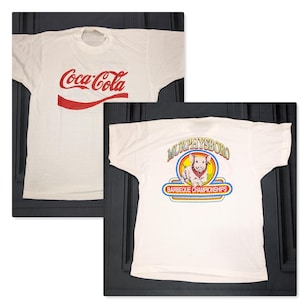 May include: Two white t-shirts. One features the Coca-Cola logo in red. The other displays a graphic of a dog wearing a bandana, with the text "Murphysboro Barbecue Championships" in a colorful design.