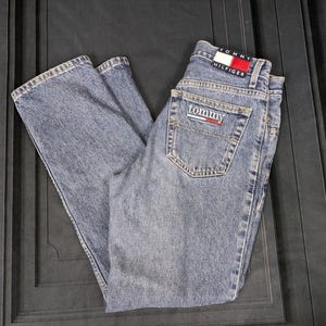 Puede incluir: Vaqueros de mezclilla azul claro con el logo "Tommy Hilfiger" en el bolsillo trasero y la cintura. Los vaqueros están doblados y parecen sin usar. Diseño clásico de cinco bolsillos.