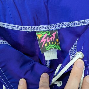Puede incluir: Primer plano de pantalones cortos de surf azules y morados con un cord&oacute;n blanco. Los pantalones cortos tienen una etiqueta que dice "Surf Style" con un dise&ntilde;o de rayo rosa. Hecho en EE. UU.