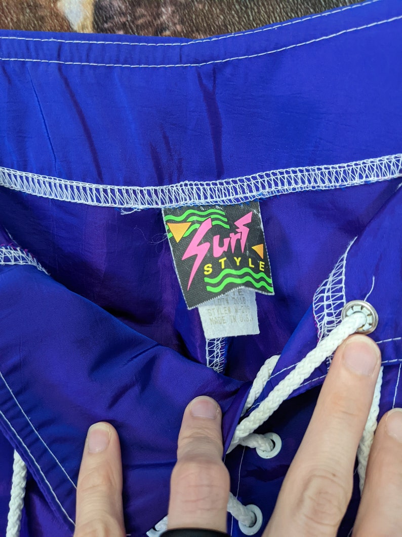 Puede incluir: Primer plano de pantalones cortos de surf azules y morados con un cord&oacute;n blanco. Los pantalones cortos tienen una etiqueta que dice "Surf Style" con un dise&ntilde;o de rayo rosa. Hecho en EE. UU.