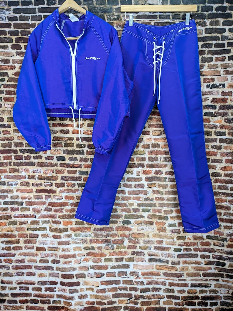 Puede incluir: Un conjunto de ch&aacute;ndal morado de dos piezas. La chaqueta tiene una cremallera y un cord&oacute;n blancos, con la palabra "Surfside" en escritura blanca. Los pantalones tienen una parte delantera con cordones. Ambas piezas tienen costuras blancas.