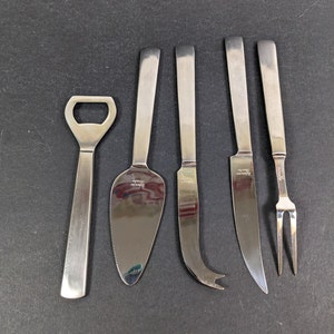 Puede incluir: Conjunto de cinco cuchillos para queso de acero inoxidable con un abridor de botellas. Los cuchillos tienen diferentes formas de hoja y todos son de color plateado.