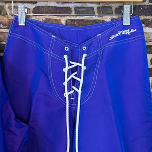 Puede incluir: Pantalones cortos de surf de color azul brillante con costuras blancas y cierre de cordones. Los pantalones cortos tienen la palabra "Surfstyle" en escritura blanca. Colgados en una percha de madera.