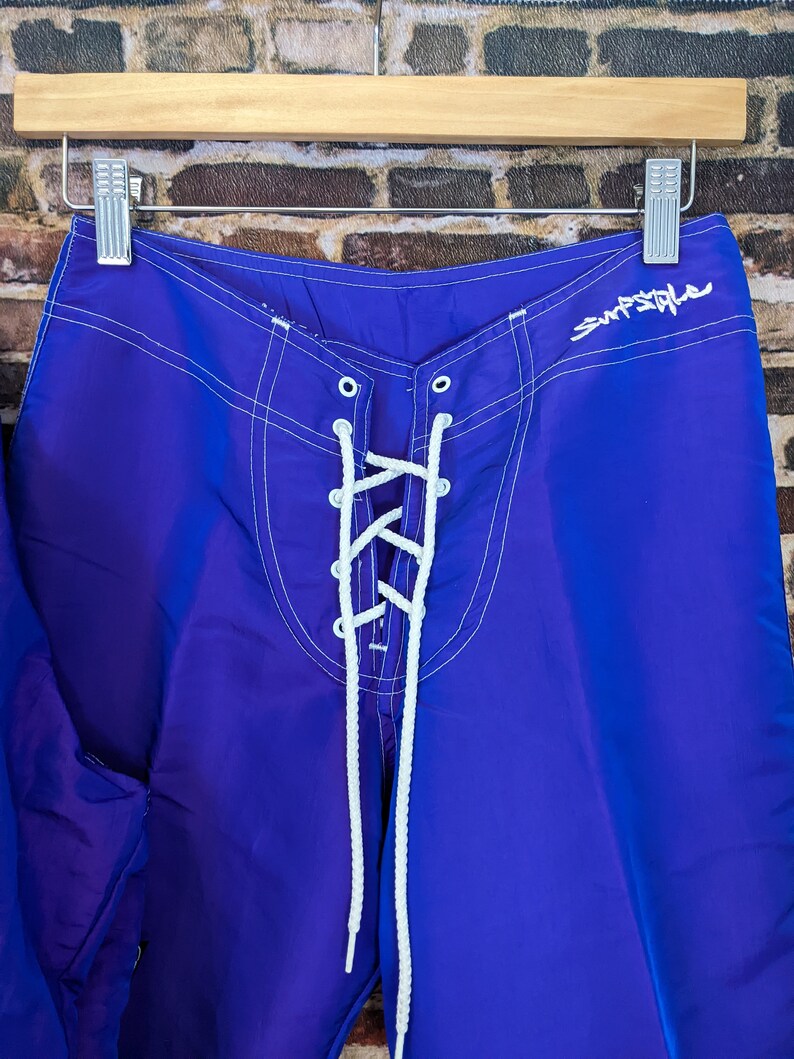 Puede incluir: Pantalones cortos de surf de color azul brillante con costuras blancas y cierre de cordones. Los pantalones cortos tienen la palabra "Surfstyle" en escritura blanca. Colgados en una percha de madera.