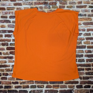May include: A sleeveless orange t-shirt. The garment is made of a soft material and is displayed against a brick wall. The shirt has a crew neck and a simple design.