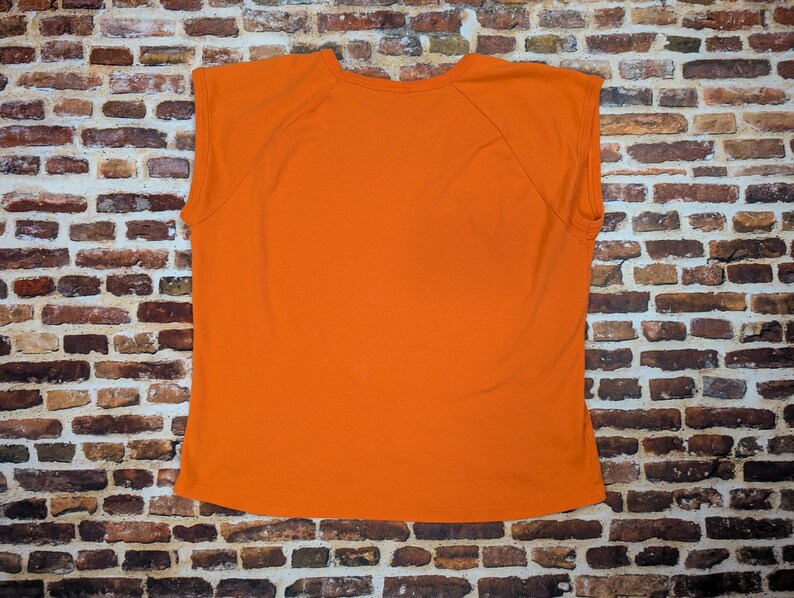 May include: A sleeveless orange t-shirt. The garment is made of a soft material and is displayed against a brick wall. The shirt has a crew neck and a simple design.
