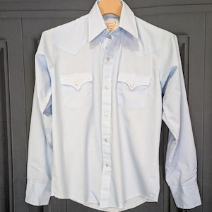 May include: A light blue Western-style shirt with long sleeves, two chest pockets, and pearl snap closures. The shirt has a pointed collar and decorative stitching on the front and back yoke. The shirt is hanging on a wooden hanger.