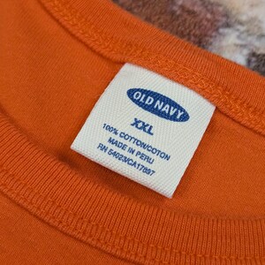 May include: Close-up of an orange Old Navy t-shirt with a white label. The label reads "OLD NAVY XXL 100% COTTON/COTON MADE IN PERU RN 54023/CA17097". The shirt has a round neckline and is made of cotton.