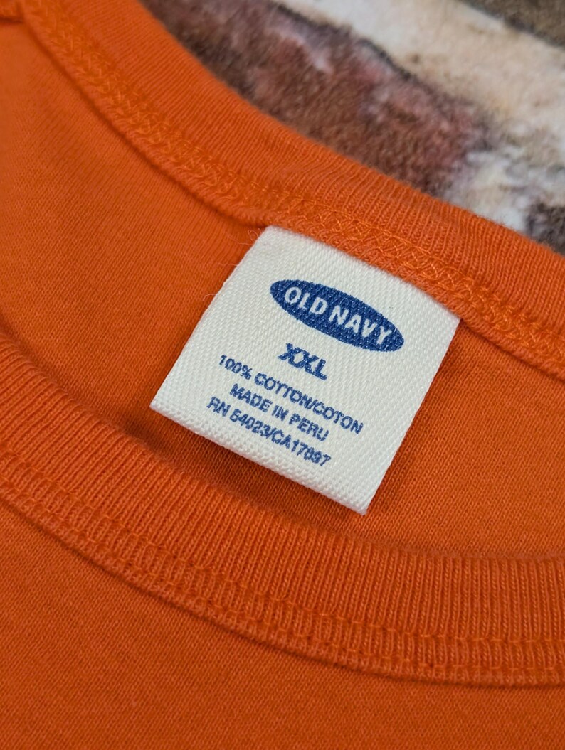 May include: Close-up of an orange Old Navy t-shirt with a white label. The label reads "OLD NAVY XXL 100% COTTON/COTON MADE IN PERU RN 54023/CA17097". The shirt has a round neckline and is made of cotton.