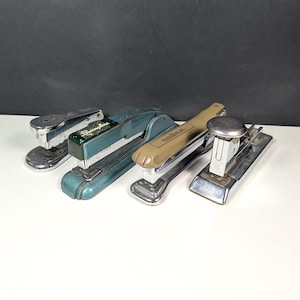 May include: Four vintage staplers in various colors, including teal, tan, and silver. The staplers are arranged on a white surface against a dark background. The brand "Swingline" is visible on one stapler.