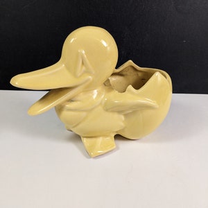 Puede incluir: Un macetero de cerámica amarillo en forma de pato con un diseño de huevo agrietado. El macetero es de estilo vintage y sería una gran adición a cualquier hogar.