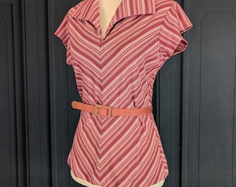 Pode incluir: Uma blusa de manga curta com decote em V, gola larga e um padrão de riscas em chevron em tons de vermelho, rosa e branco. A peça é ajustada na cintura com um cinto fino, texturizado e de cor sólida com uma fivela retangular.