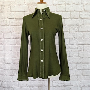 May include: Olive green long-sleeve shirt with a pointed collar and white buttons. The shirt has a fitted silhouette and is displayed on a mannequin. The stitching is white, providing contrast to the dark green fabric.