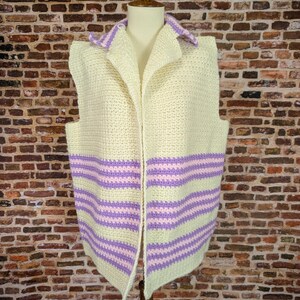 May include: A cream-colored, open-front crochet vest with a collar and horizontal stripes in lavender and light pink. The vest is displayed on a mannequin against a brick wall background.